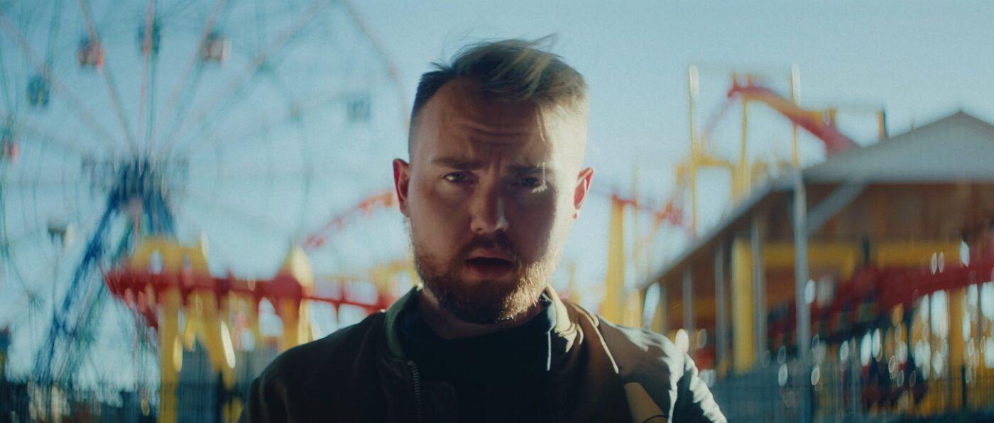 Trey Abdella with a beard stands outdoors in front of an amusement park with a Ferris wheel and colorful rides visible in the background.