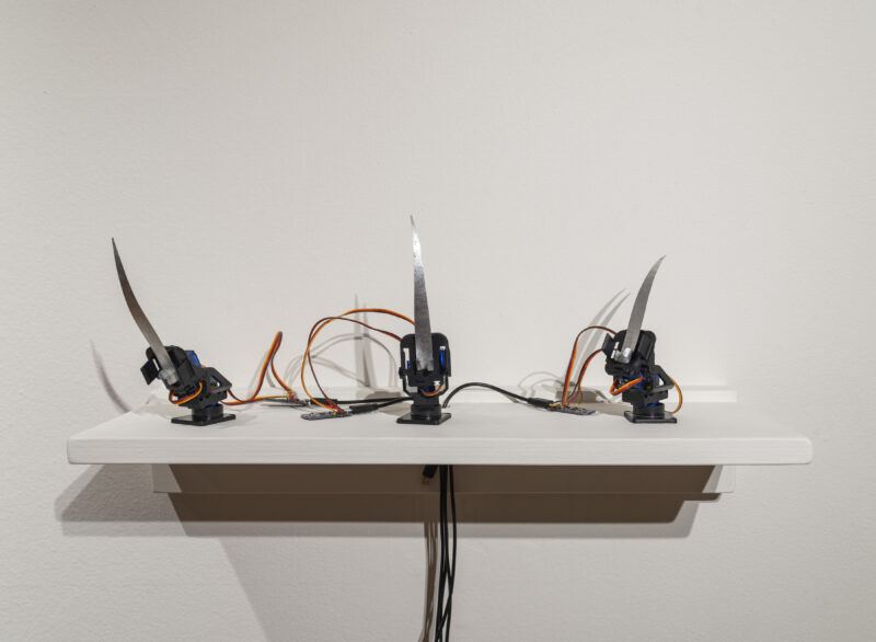Three robotic arms with attached knives are mounted on a white shelf against a plain wall, with wires visible connecting the devices.