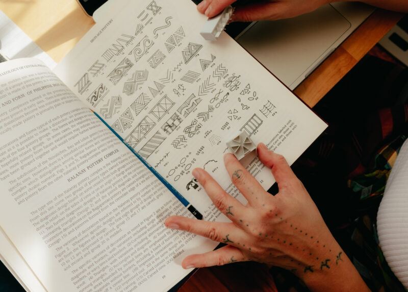 A person holding a book showing various geometric patterns and symbols, with their tattooed hand holding a small object next to the open pages.