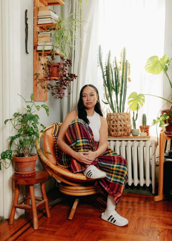Caroline Garcia in a white sleeveless top and plaid skirt sits on a chair surrounded by various potted plants in a sunlit room.