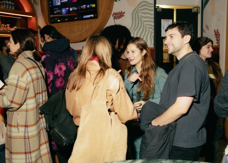 A group of people socialize indoors at what appears to be a lively gathering, with some holding drinks and chatting.