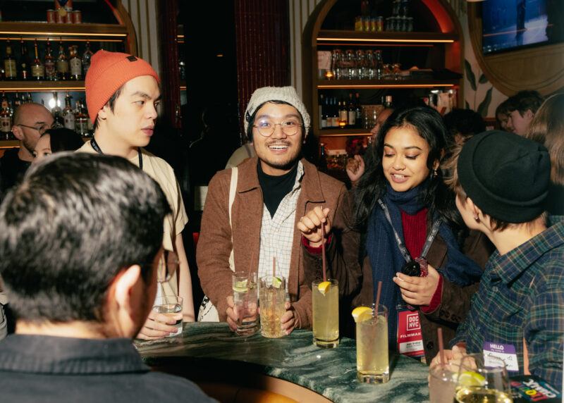 A group of people stand around a bar table with drinks, conversing and smiling in a dimly lit bar.