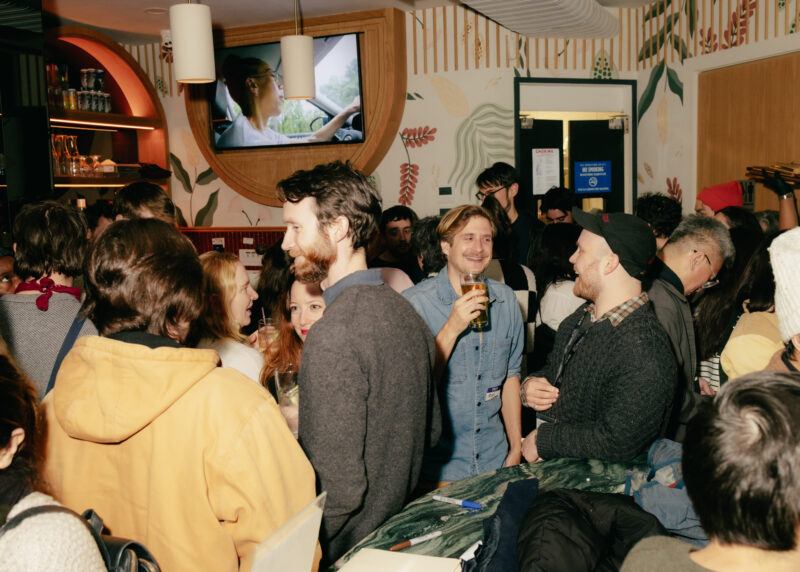 A crowded bar scene with people socializing, some holding drinks, and a TV in the background displaying a woman driving a car.