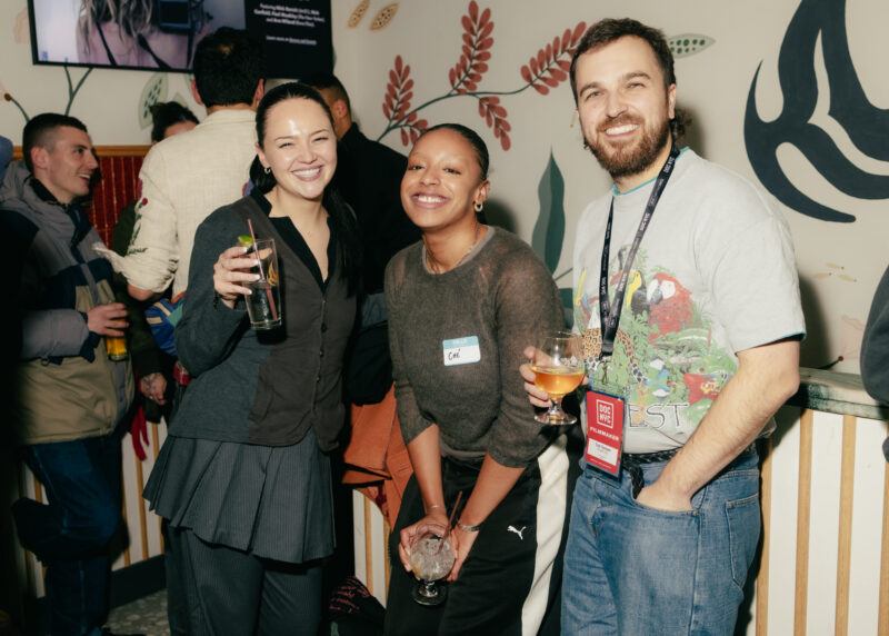 Three people stand together at an indoor social event, smiling at the camera while holding drinks; other attendees are visible in the background.