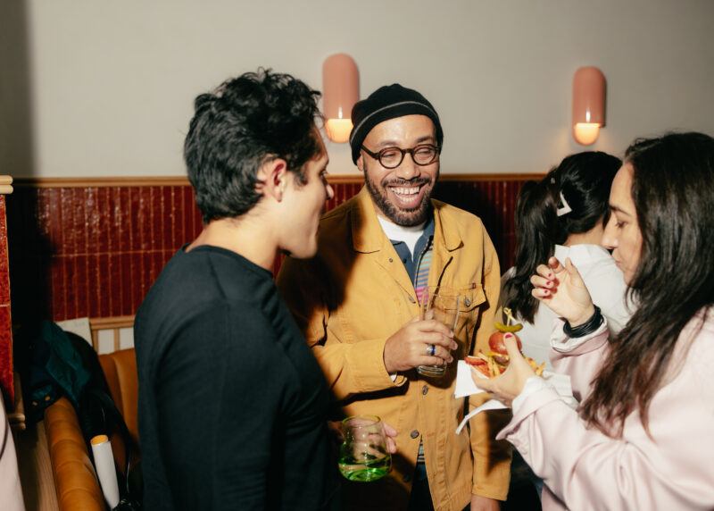 Three people socialize indoors at a gathering; one man in glasses and a beanie smiles while holding a drink, others are eating and talking.