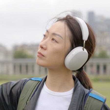 Xin Liu wearing large white headphones and a backpack looks upward while standing outdoors on a cloudy day.